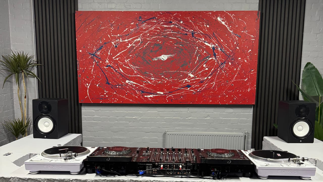 The DJ room square-on: two AlphaTheta CDJ-3000X players either side of a Pioneer DJ DJM-A9 with a white Reloop RP-7000 turntable at each end, Yamaha HS8 monitors on the wings of a hand-built concrete booth, beneath a red and white abstract canvas