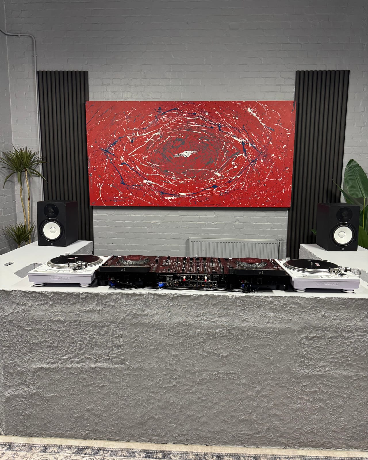 The DJ room in full: the red and white abstract canvas above a hand-built concrete booth carrying two AlphaTheta CDJ-3000X players, a Pioneer DJ DJM-A9 and two white Reloop RP-7000 turntables, with Yamaha HS8 monitors on the booth's concrete wings