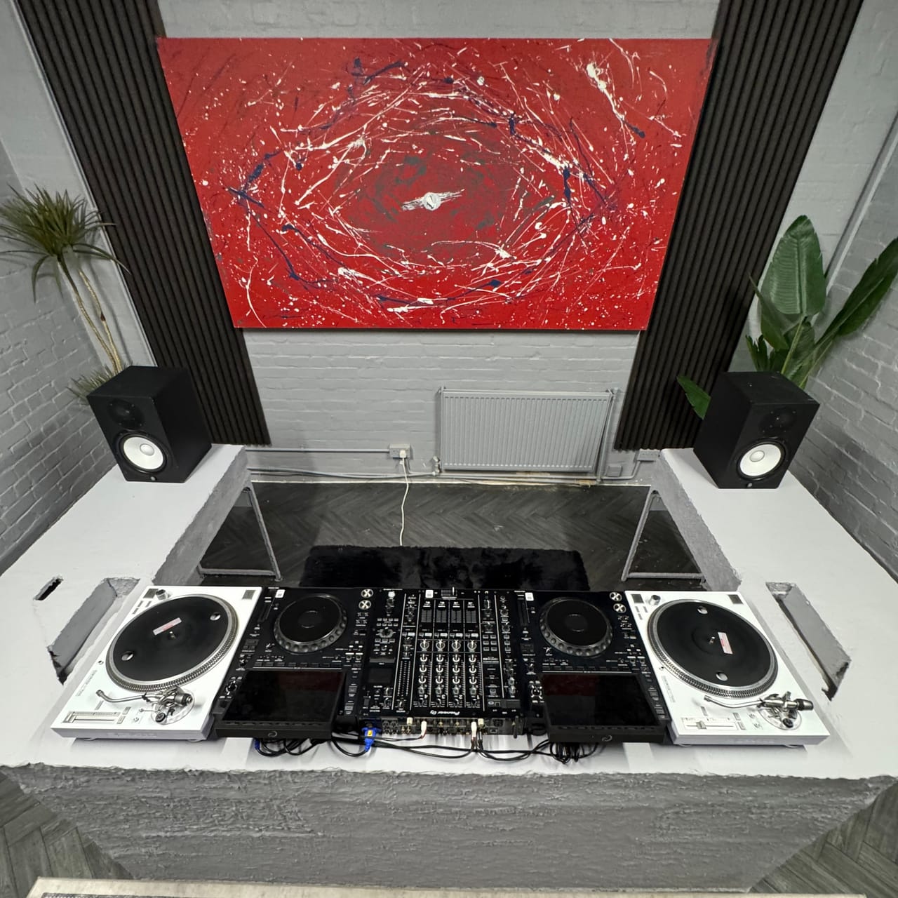 The DJ booth square-on from the DJ's side: two white Reloop RP-7000 turntables at the ends, two AlphaTheta CDJ-3000X players and a Pioneer DJ DJM-A9 across the middle, Yamaha HS8 monitors on the concrete wings and the red and white abstract canvas whole on the wall above