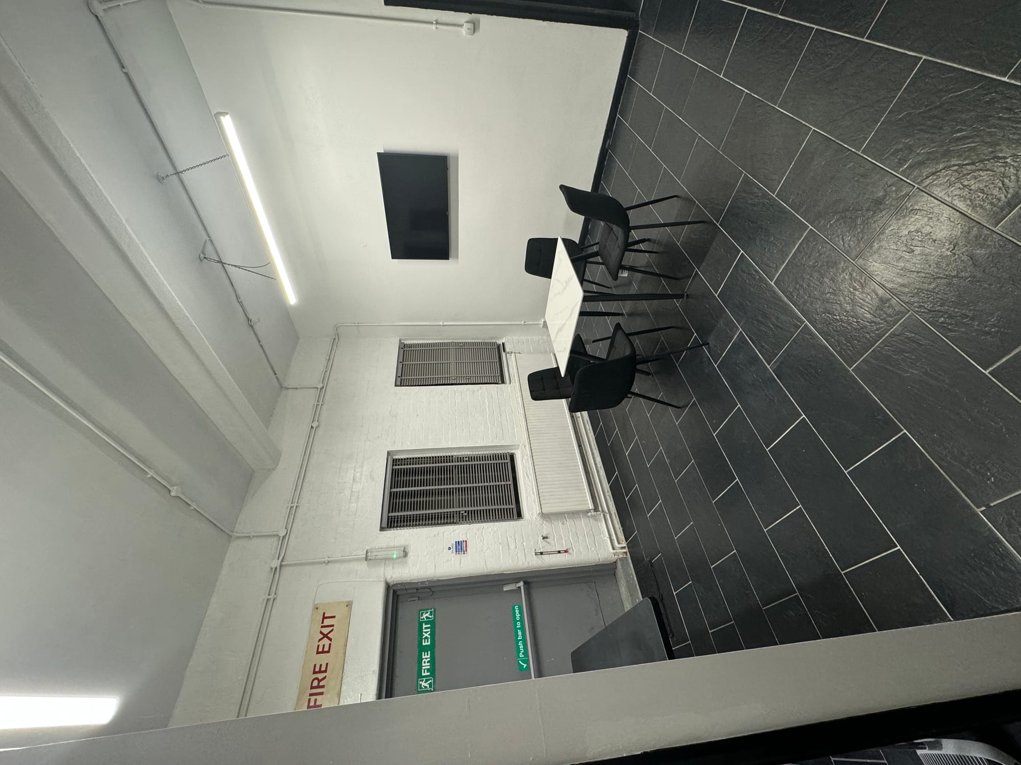 The kitchen: a white table with four black chairs on a slate tiled floor, with a wall-mounted television and a fire exit