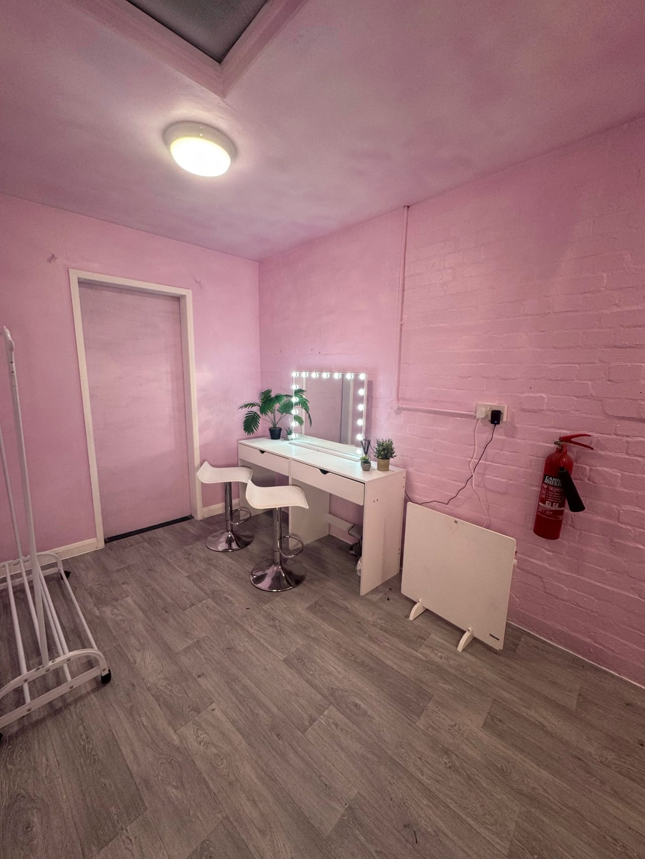 The prep and dressing room: pink painted walls, a white vanity desk with a bulb-surround mirror, two stools and a clothes rail