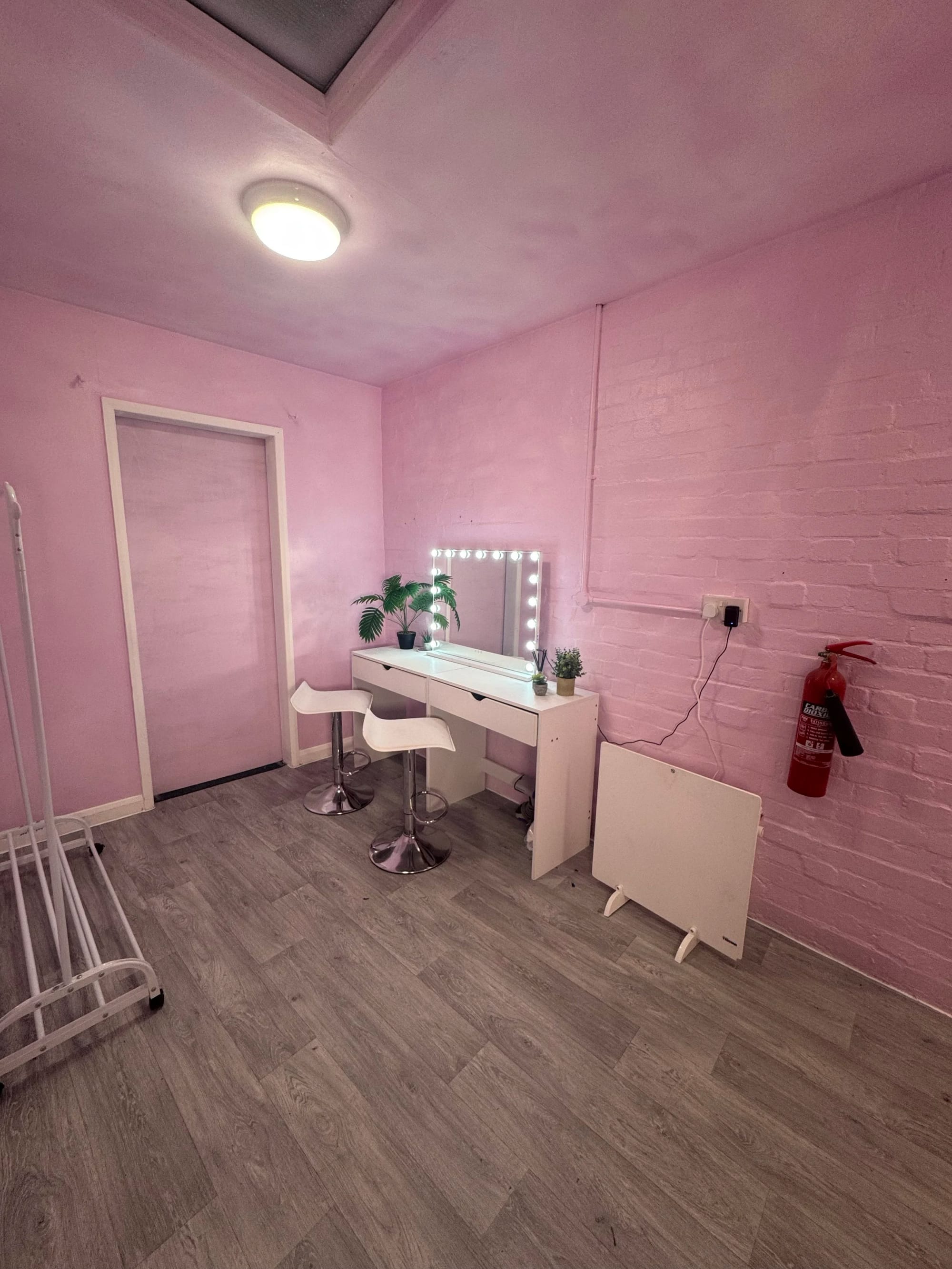 The prep and dressing room: pink painted walls, a white vanity desk with a bulb-surround mirror, two stools and a clothes rail