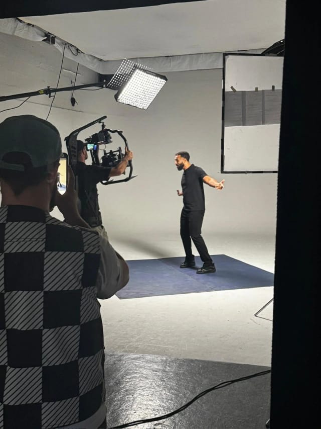 Behind the scenes in Studio A: a presenter on a blue floor mat lit by an LED panel and a poly bounce