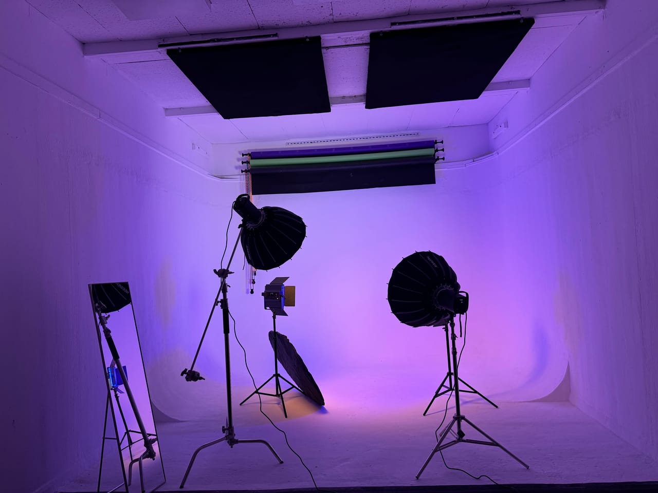 Studio B's white cyclorama washed in deep purple light, with two large para softboxes, an LED panel, a reflector and a standing mirror arranged across the cove, and coloured backdrop rolls stowed above