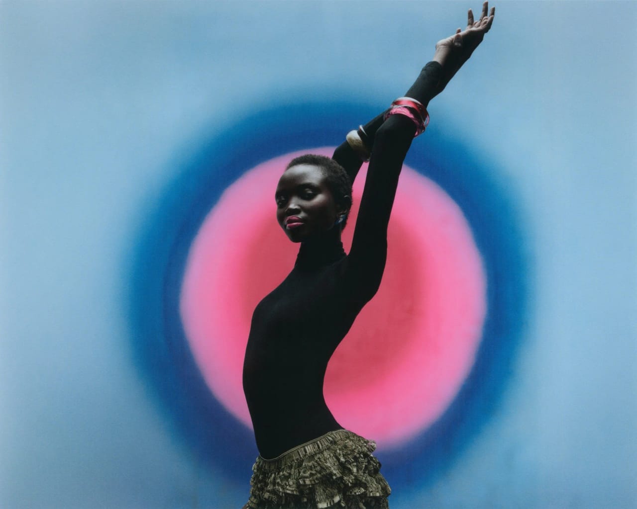 Fashion editorial in landscape: a model in a black top and gold skirt with one arm raised against a blue and pink painted backdrop