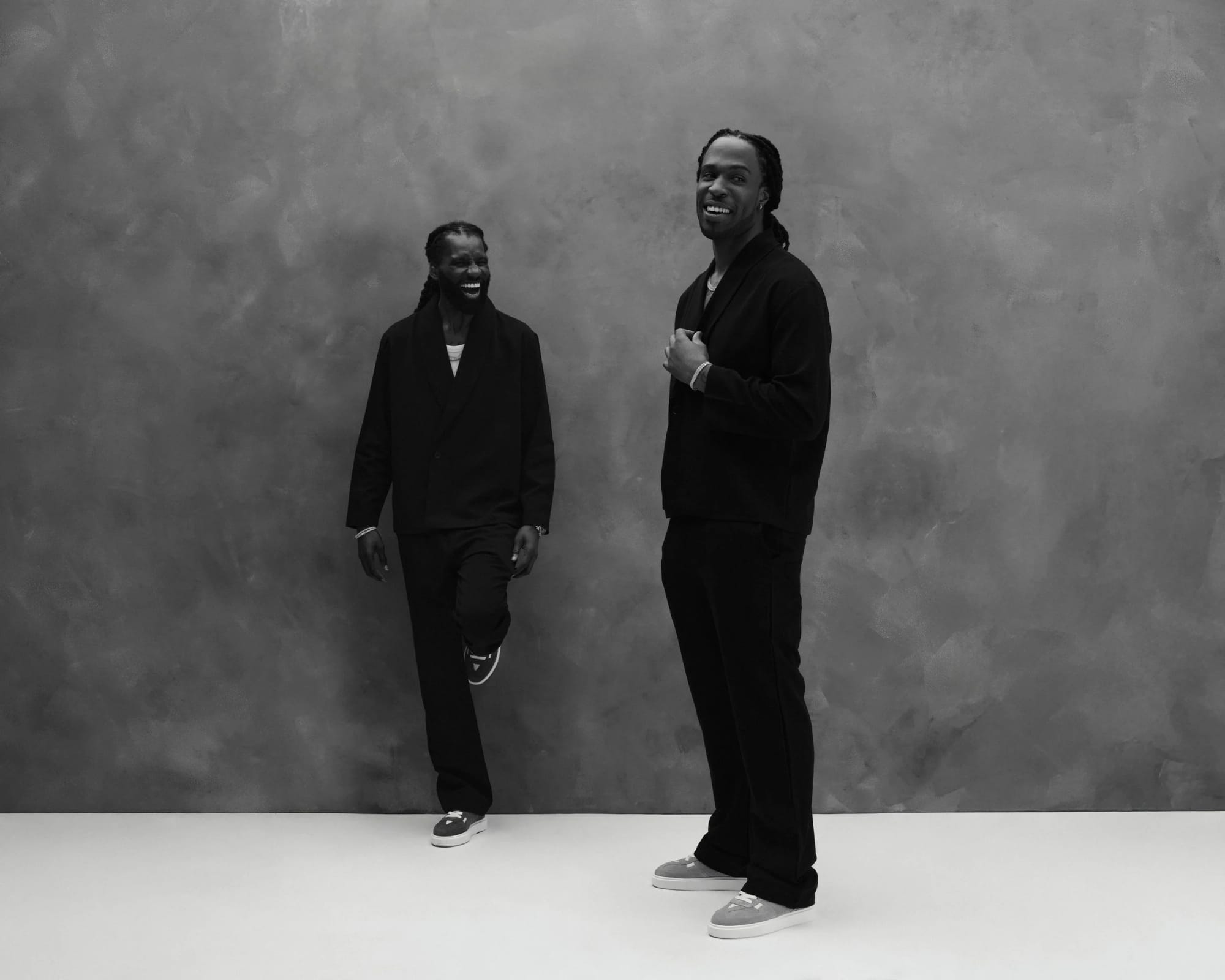 Two artists photographed together in black on a mottled grey backdrop, mid-laugh